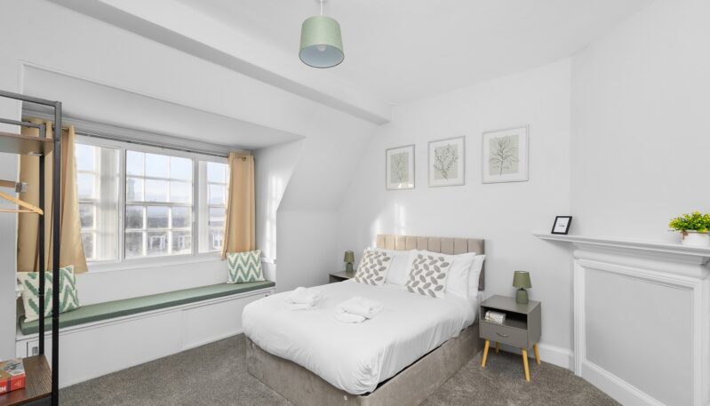 A contemporary bedroom with white walls, grey carpeting, and a double bed with a grey velvet headboard. A large bay window includes a built-in bench with green cushions, and three botanical art prints hang above the bed.