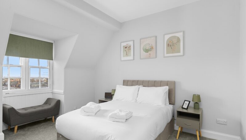 A second view of a modern bedroom showing a double bed with white linens and rolled towels. To the left, a grey upholstered chaise longue sits beneath a window with a green roller blind, and two grey nightstands with green lamps flank the bed.