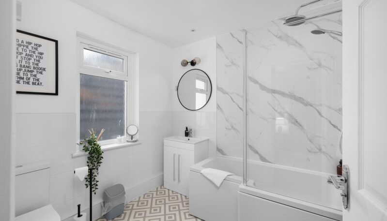 A bright, contemporary bathroom with geometric patterned floor tiles and white wall tiling. The room includes a white vanity unit with a circular mirror, a toilet, and a bathtub with a glass screen and marble-patterned wet wall panels.