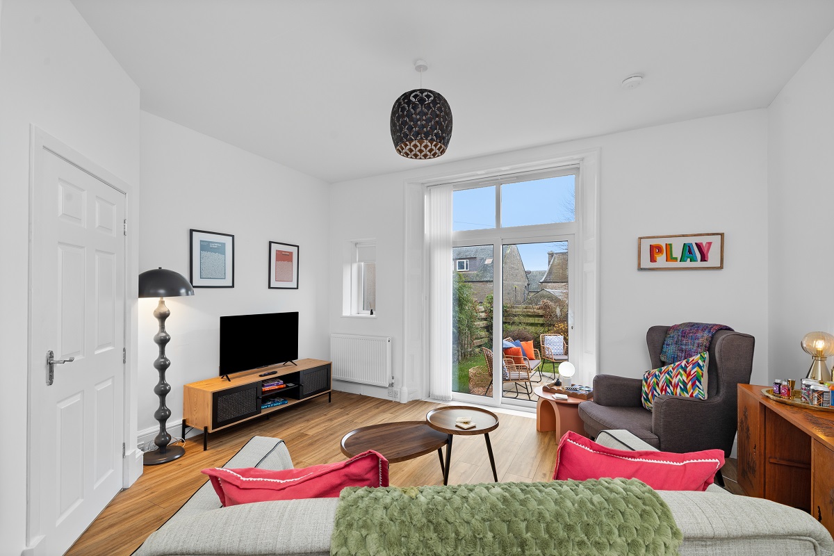 A vibrant living room featuring wood floors and a large sliding glass door that leads to a patio. The space is furnished with a grey armchair, a colorful "PLAY" wall sign, a flat-screen TV on a wooden stand, and a grey sofa with bright pink cushions.