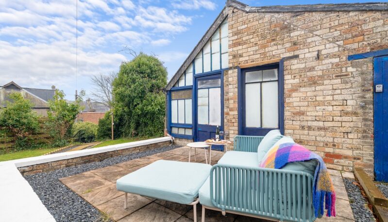 An outdoor patio area with stone paving and a small gravel border next to a rustic brick building with blue-trimmed windows. A modern light-blue outdoor sofa with a matching ottoman and a colorful plaid blanket sits in the foreground.