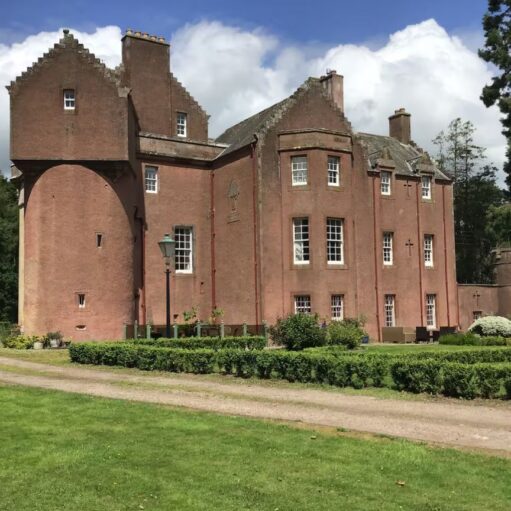 A large, historic Scottish baronial mansion with a pinkish-red sandstone exterior under a bright, partly cloudy sky. The building features distinctive crow-stepped gables, various tower wings, and small sash windows. A manicured green lawn with a low hedge and a gravel driveway sits in the foreground, flanked by tall green trees.