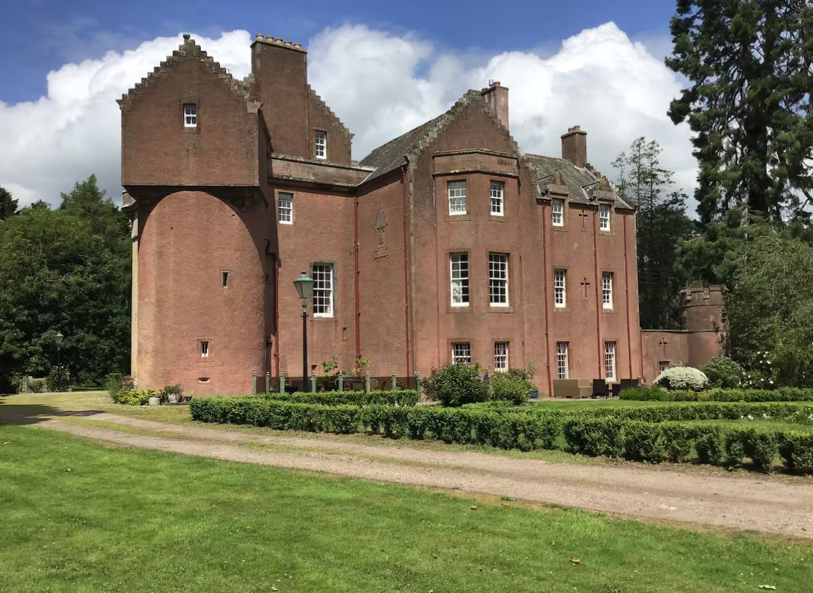 A large, historic Scottish baronial mansion with a pinkish-red sandstone exterior under a bright, partly cloudy sky. The building features distinctive crow-stepped gables, various tower wings, and small sash windows. A manicured green lawn with a low hedge and a gravel driveway sits in the foreground, flanked by tall green trees.