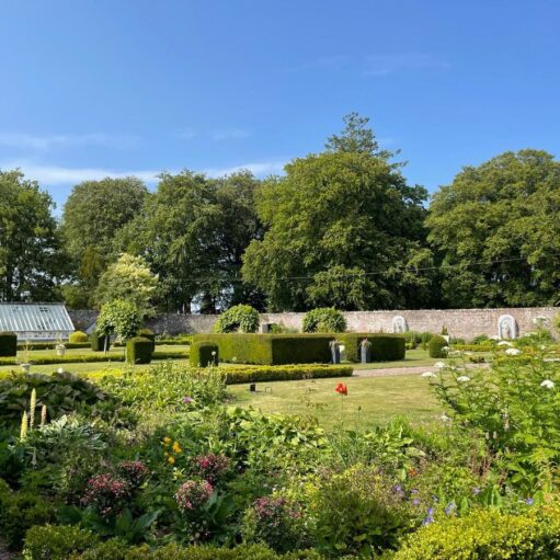A vibrant, sunlit formal garden featuring meticulously manicured box hedges, green lawns, and colorful flower beds. In the background, a long stone wall separates the garden from a dense line of tall, leafy trees under a clear blue sky, with a small glass greenhouse visible on the left.