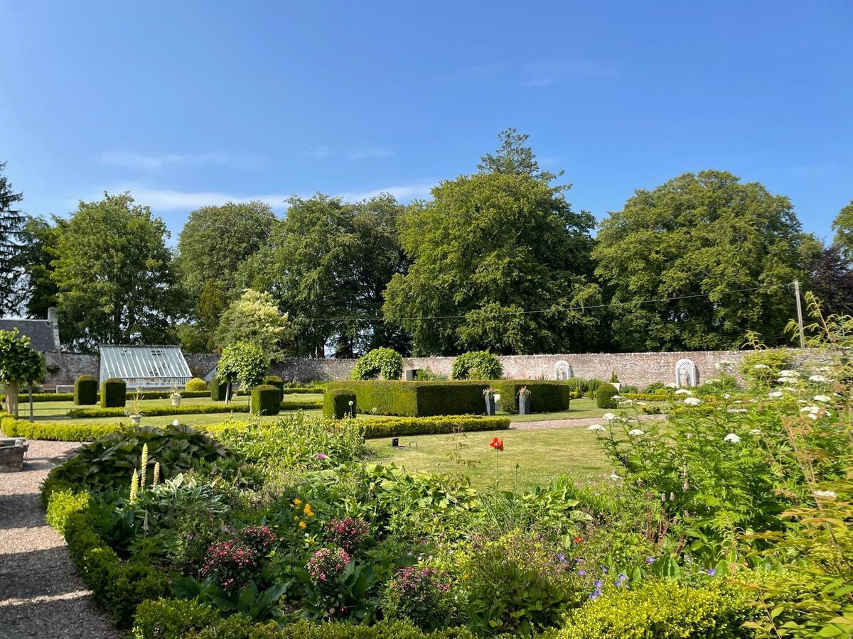 A vibrant, sunlit formal garden featuring meticulously manicured box hedges, green lawns, and colorful flower beds. In the background, a long stone wall separates the garden from a dense line of tall, leafy trees under a clear blue sky, with a small glass greenhouse visible on the left.