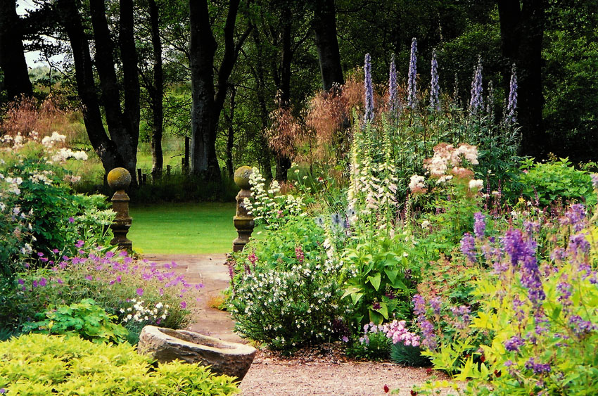 A gravel and stone pathway winds through a lush, colorful garden filled with purple delphiniums, pink flowers, and various green shrubs. In the background, two stone bollards mark an entrance to a bright green lawn, framed by a dense stand of dark trees.