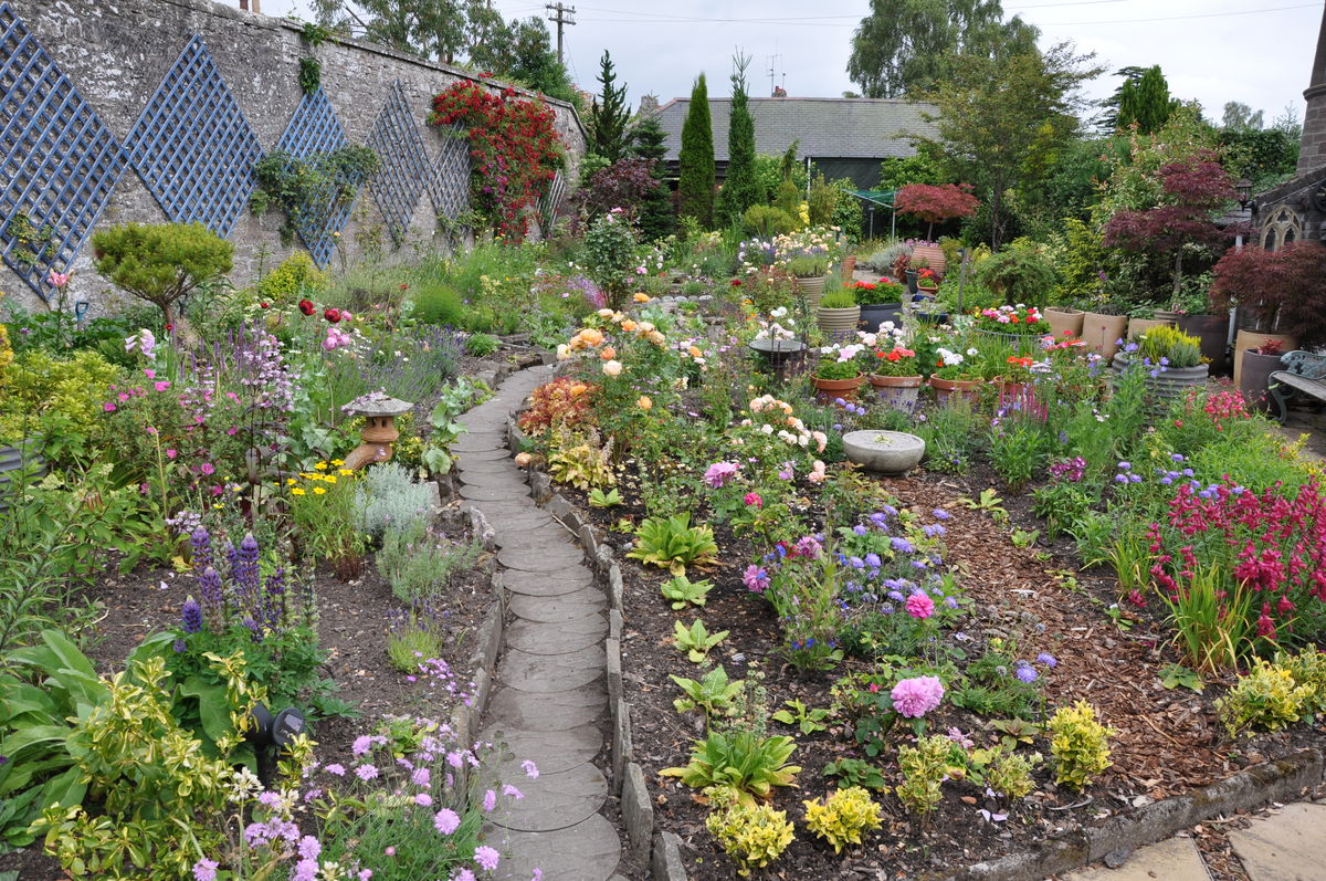 A vibrant, overflowing walled garden featuring a narrow, winding stone path made of circular pavers. The garden is filled with a variety of colorful flowers including purple lupines, pink roses, and yellow wildflowers, interspersed with potted plants and garden ornaments. A tall stone wall on the left is decorated with blue diamond-shaped trellises and climbing red roses.