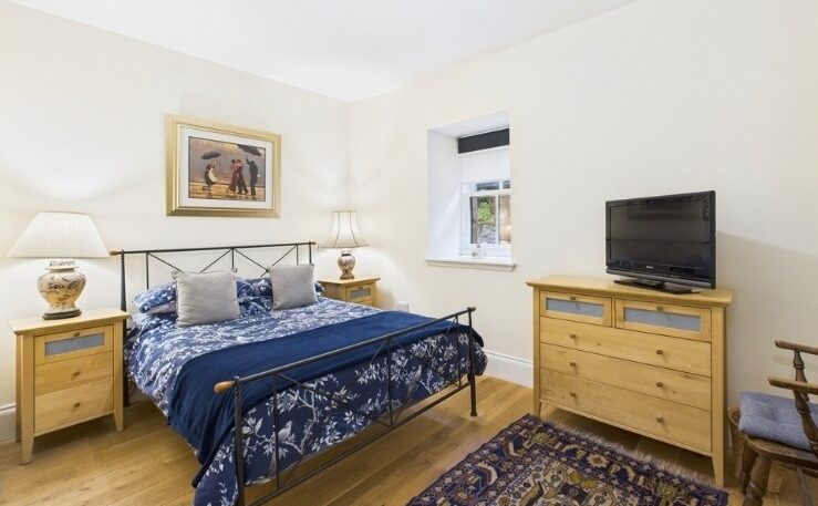 A bright and airy bedroom with a double bed dressed in blue floral linens and a navy throw. The room includes light wood bedside tables with matching lamps, a chest of drawers with a television, and a framed painting hanging above the black metal headboard.