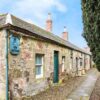 A long, traditional stone cottage with a slate roof and green doors. A blue sign reading "Yew Cottage" is mounted on the corner of the rugged stone wall, and a stone-paved path runs alongside the building next to a tall, manicured hedge.