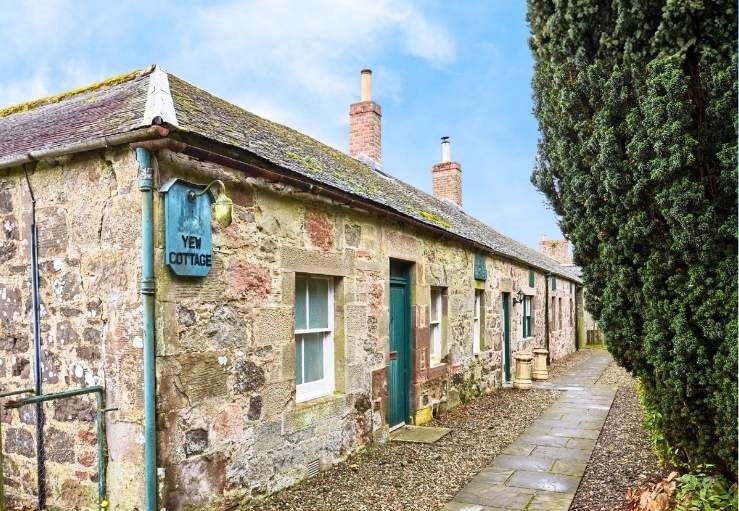 A long, traditional stone cottage with a slate roof and green doors. A blue sign reading "Yew Cottage" is mounted on the corner of the rugged stone wall, and a stone-paved path runs alongside the building next to a tall, manicured hedge.