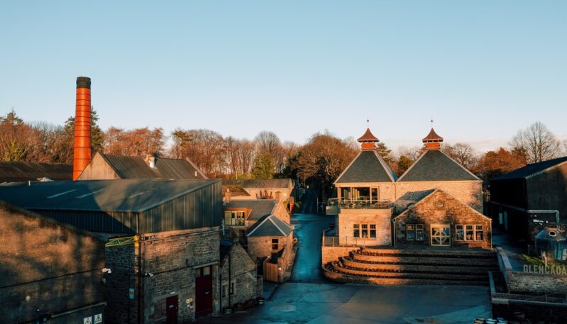 An elevated drone view of the Glencadam Distillery during golden hour, featuring historic stone buildings, traditional pagoda-style kiln roofs, and a tall red-brick chimney set against a backdrop of trees.