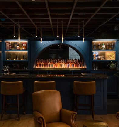 An elegant, dimly lit interior bar featuring deep blue wood-paneled walls, leather armchairs, a marble-top bar, and back-lit shelves displaying various whisky bottles.