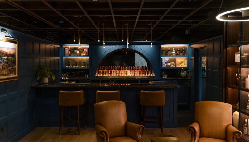 An elegant, dimly lit interior bar featuring deep blue wood-paneled walls, leather armchairs, a marble-top bar, and back-lit shelves displaying various whisky bottles.