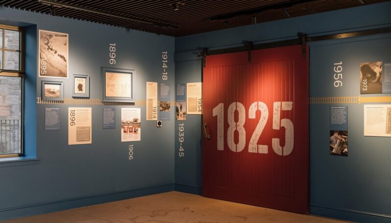 A historical timeline wall in the visitor center painted deep blue, decorated with dates, vintage photos, and a large sliding red barn door with "1825" printed on it in white.