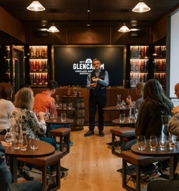 A guided whisky tasting session taking place in a modern room, where a presenter stands at the front while guests sit at small tables with Glencairn glasses and water carafes.