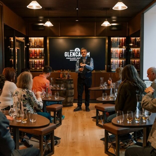 A guided whisky tasting session taking place in a modern room, where a presenter stands at the front while guests sit at small tables with Glencairn glasses and water carafes.