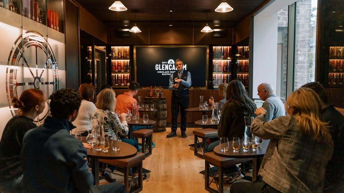 A guided whisky tasting session taking place in a modern room, where a presenter stands at the front while guests sit at small tables with Glencairn glasses and water carafes.