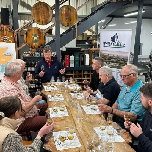 A group of people participate in a whisky tasting session around a long, natural-edge wooden table in a rustic distillery setting. A presenter at the head of the table gestures while holding a tasting glass, while participants observe and sample various spirits. The background features decorative barrel heads, a map of Scotland, and a "Whisky Caddie" banner.