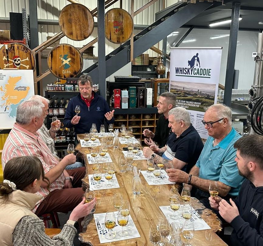 A group of people participate in a whisky tasting session around a long, natural-edge wooden table in a rustic distillery setting. A presenter at the head of the table gestures while holding a tasting glass, while participants observe and sample various spirits. The background features decorative barrel heads, a map of Scotland, and a "Whisky Caddie" banner.