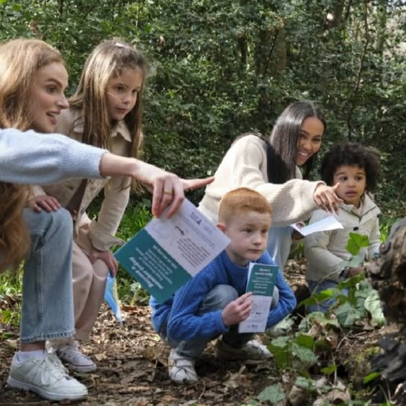 Two women and three children are exploring a wooded area together. One woman and child point toward something off-camera while the others look on curiously. The group is holding informational pamphlets as they crouch along a leafy forest path.