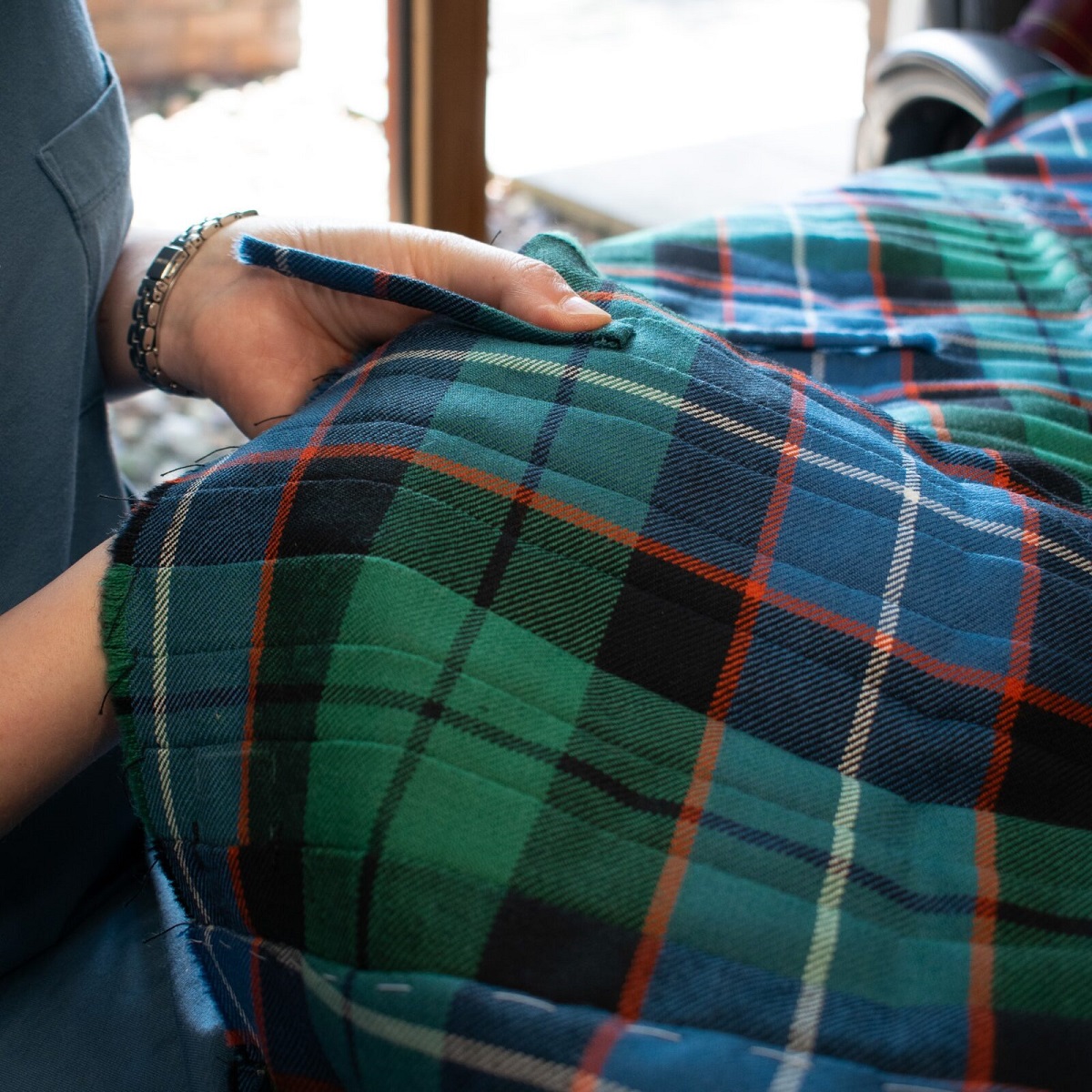 A close-up photograph shows a person's hands meticulously working on a traditional green, blue, and red tartan kilt. The fabric is thick and textured, featuring visible white stitching used for marking or basting. The artisan is shown handling the heavy material near a window, emphasizing the detailed craftsmanship involved in Scottish tailoring.