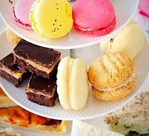 A three-tiered white cake stand is filled with a variety of afternoon tea treats. The top tier features brightly colored pink and yellow macarons, the middle tier contains chocolate brownies and cream-filled cookies, and the bottom tier holds savory finger sandwiches and slices of quiche. Pink tulips are visible in the blurred background.