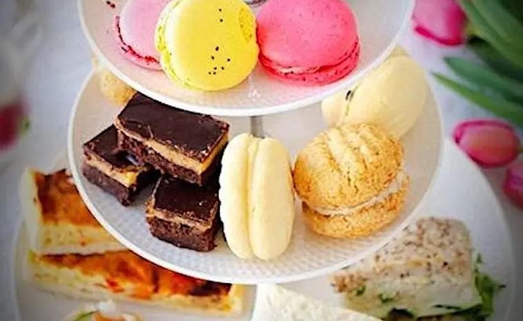 A three-tiered white cake stand is filled with a variety of afternoon tea treats. The top tier features brightly colored pink and yellow macarons, the middle tier contains chocolate brownies and cream-filled cookies, and the bottom tier holds savory finger sandwiches and slices of quiche. Pink tulips are visible in the blurred background.