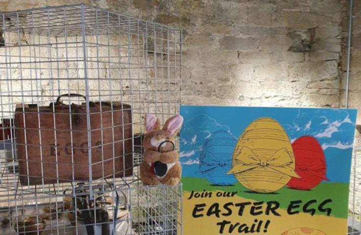 A plush Easter bunny sits on a wire mesh cage next to a colorful sign that says, "Join our EASTER EGG Trail!" Inside the cage is a vintage wooden crate labeled "EGGS." The sign features illustrations of blue, yellow, and red Easter eggs with ribbons against a blue sky.