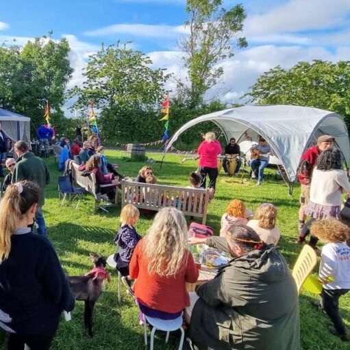 Gemini said A wide shot of an outdoor community gathering on a grassy field under a bright, partly cloudy sky. People of various ages are seated on wooden benches and folding chairs or standing around, many with drinks. In the background, a musical duo performs under a gray canopy tent, while colorful rainbow flags and bunting decorate the area.