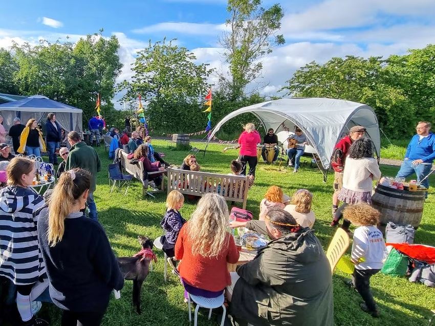 Gemini said A wide shot of an outdoor community gathering on a grassy field under a bright, partly cloudy sky. People of various ages are seated on wooden benches and folding chairs or standing around, many with drinks. In the background, a musical duo performs under a gray canopy tent, while colorful rainbow flags and bunting decorate the area.