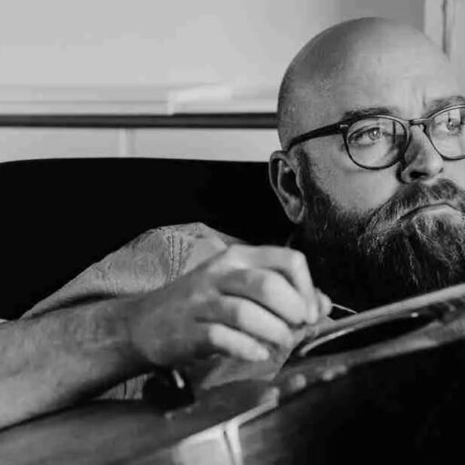 A black-and-white, close-up photograph of a man with a thick beard and glasses, looking thoughtfully off to the side. He is shown from the chest up, leaning forward slightly as he works on or holds a musical instrument, likely a guitar.
