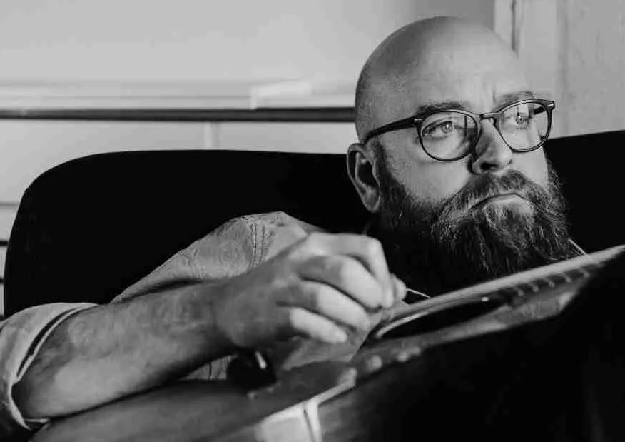 A black-and-white, close-up photograph of a man with a thick beard and glasses, looking thoughtfully off to the side. He is shown from the chest up, leaning forward slightly as he works on or holds a musical instrument, likely a guitar.