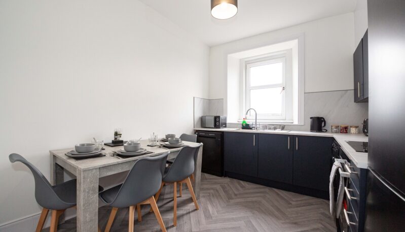 A modern dining kitchen featuring a grey wood-effect table set for four, grey bucket chairs, and a herringbone-patterned floor. The kitchen area has dark navy cabinetry, white countertops, and integrated appliances under a bright window.