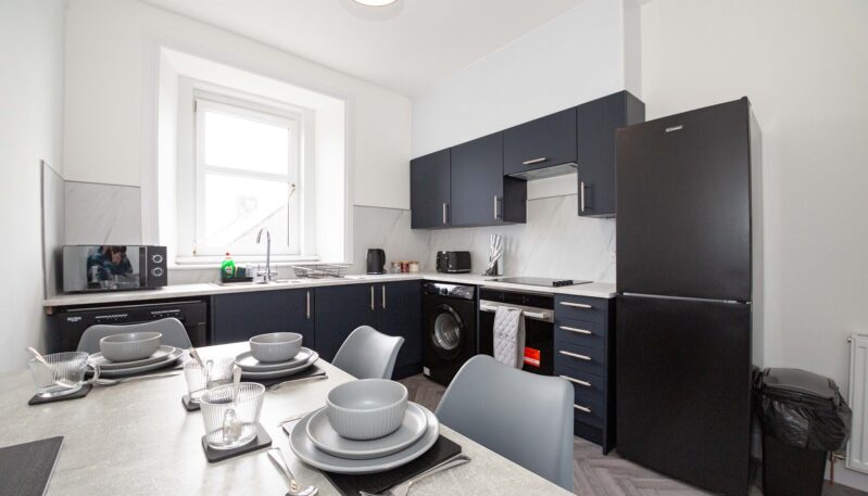 A bright kitchen showing a full suite of navy blue cabinets and white worktops. It includes a black fridge-freezer, an integrated oven and hob, a washing machine, and a microwave, with a dining table in the foreground.