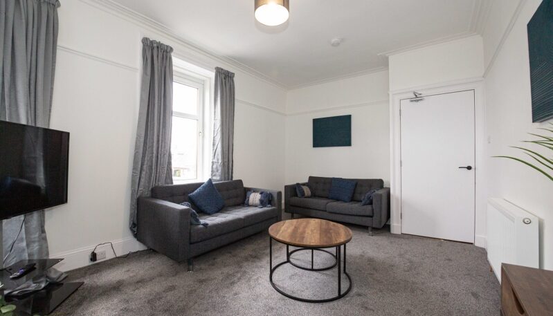 A bright living area with two grey fabric sofas adorned with blue cushions. A round wooden coffee table sits in the center of the room on a dark grey carpet, positioned near a large window with grey curtains.