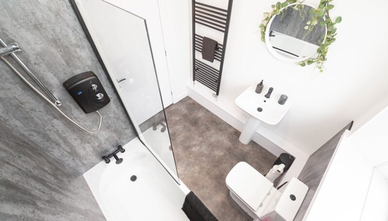 A high-angle view of a contemporary bathroom with grey marble-effect wall panels. It features a white bathtub with an overhead electric shower, a pedestal sink, a toilet, and a black heated towel rail.