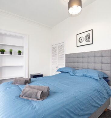 A minimalist bedroom with a grey upholstered headboard and a bed made with blue linens and folded grey towels. The room includes white walls, a built-in shelving unit with small plants, and a modern pendant light.