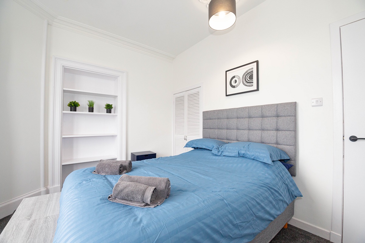 A minimalist bedroom with a grey upholstered headboard and a bed made with blue linens and folded grey towels. The room includes white walls, a built-in shelving unit with small plants, and a modern pendant light.