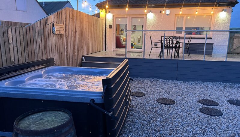 A modern backyard patio at dusk featuring a hot tub, a raised composite deck with outdoor seating, and warm string lights overhead.