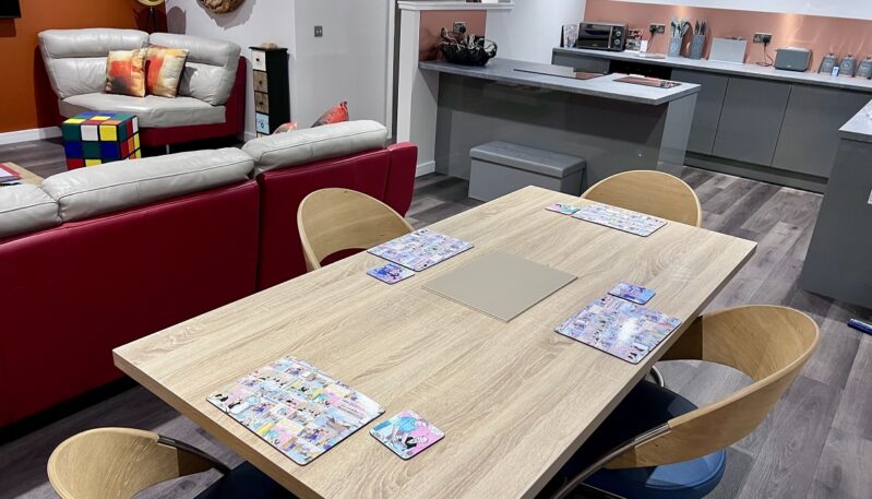 Inside a modern open-plan home showing a dining area with a wooden table, overlooking a kitchen with gray cabinets and a living room with a red sofa.