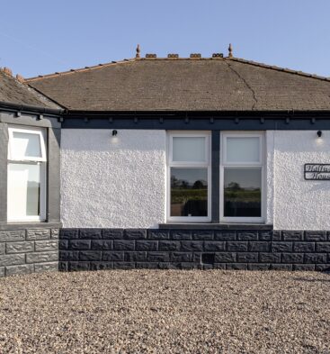 Exterior view of a modern single-story bungalow with white walls, dark gray stone detailing, and a gravel front garden.