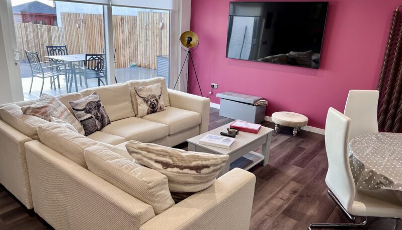 A cosy living room with a large comfortable sofa, a pink accent wall with a mounted television, and a sliding glass door leading to a patio.