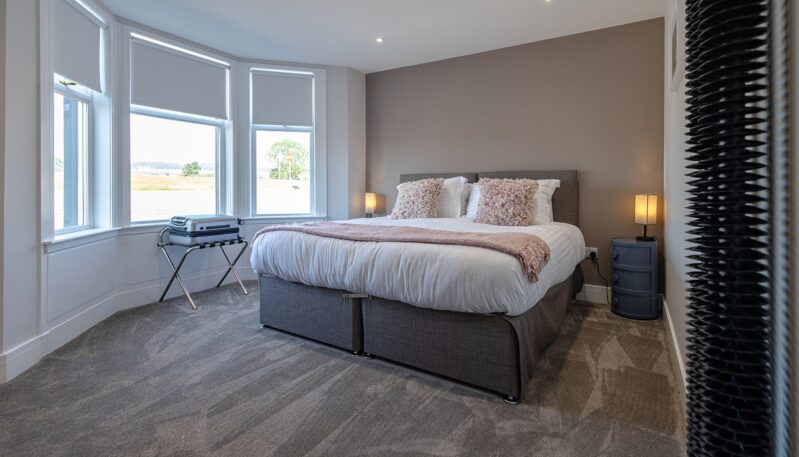 A modern, minimalist bedroom with a large bed, neutral-toned walls, and a bay window overlooking a rural landscape.