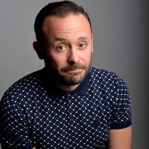 A studio headshot of a man with short dark hair and a groomed beard, looking at the camera with a playful, slightly raised eyebrow and a wry expression. He is wearing a dark blue polo shirt featuring a small, white geometric pattern. The background is a simple, neutral grey gradient.