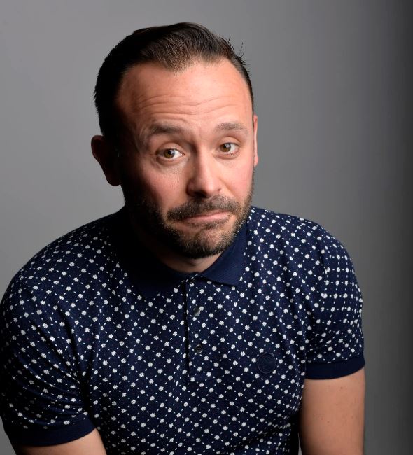 A studio headshot of a man with short dark hair and a groomed beard, looking at the camera with a playful, slightly raised eyebrow and a wry expression. He is wearing a dark blue polo shirt featuring a small, white geometric pattern. The background is a simple, neutral grey gradient.