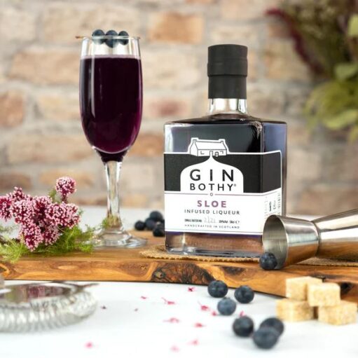 A bottle of "Gin Bothy" sloe infused liqueur sits next to a glass filled with the dark purple liquid, placed on a wooden board with scattered blueberries, sugar cubes, and small pink flowers against a rustic brick background.