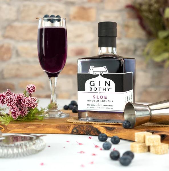 A bottle of "Gin Bothy" sloe infused liqueur sits next to a glass filled with the dark purple liquid, placed on a wooden board with scattered blueberries, sugar cubes, and small pink flowers against a rustic brick background.