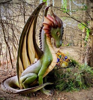 A detailed statue of a green dragon with large wings sits in a wooded area, guarding a small pile of treasure partially hidden behind bushes.