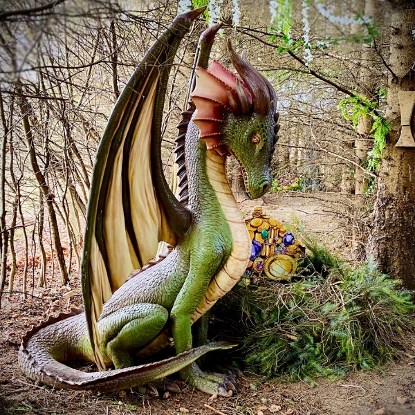 A detailed statue of a green dragon with large wings sits in a wooded area, guarding a small pile of treasure partially hidden behind bushes.