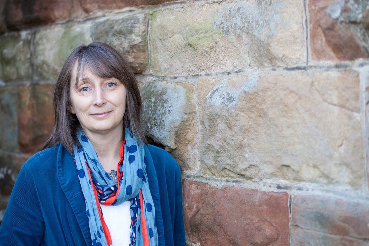 An image of a woman with medium-length brown hair and bangs, smiling gently while leaning against a rustic stone wall. She is wearing a blue jacket and a patterned light blue scarf with dark blue circles and a thin red border.